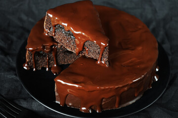 Chocolate cake with chocolate icing on a black plate next to a fork on a dark background