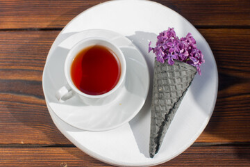red tea in a mug on a saucer with a waffle black horn and a bouquet of lilacs. Summer hot drink