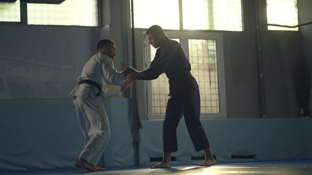 Young caucasian man in blue kimono throwing his opponent on floor while practicing jujitsu takedown in gym