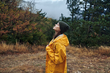 A contemplative woman in a vibrant yellow raincoat gazes upward at the sky against a backdrop of a...