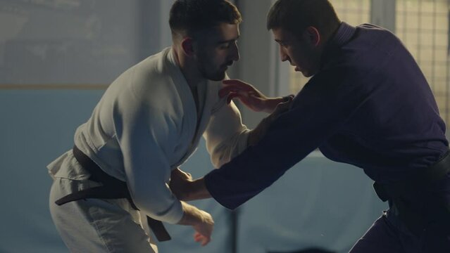 Slow-motion shot of barefoot martial arts athlete in blue kimono lifting his opponent up and throwing him down on mat during jujitsu practice in gym