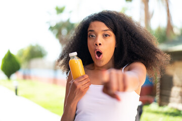 Young African American woman holding an orange juice at outdoors surprised and pointing front