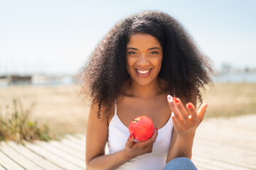 Young African American woman with an apple at outdoors inviting to come with hand. Happy that you...