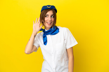 Airplane stewardess woman isolated on yellow background listening to something by putting hand on the ear