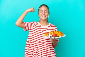 Little caucasian girl holding waffles isolated on blue background doing strong gesture