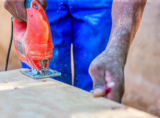 african man using a jigsaw to cut a plank of wood, holding a pencil to mark
