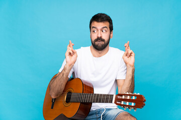 Young man with guitar over isolated blue background with fingers crossing and wishing the best