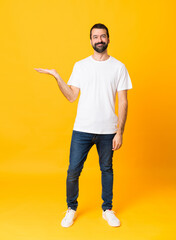 Full-length shot of man with beard over isolated yellow background holding copyspace imaginary on...