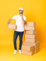 Full-length shot of delivery man among boxes over isolated yellow background showing a sign of...