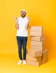 Full-length shot of delivery man among boxes over isolated yellow background with fingers crossing