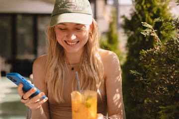 Happy laughing blonde woman in cap sitting in cafe and drink cocktail. Girl hold mobile phone read...