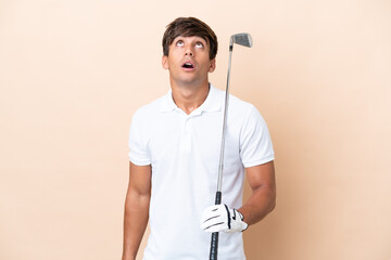 Young golfer player man isolated on ocher background looking up and with surprised expression