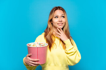 Teenager girl holding popcorns over isolated blue background looking up while smiling