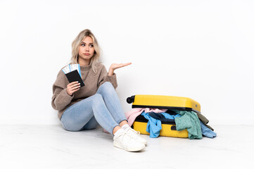 Teenager blonde girl with a suitcase full of clothes sitting on the floor holding copyspace with doubts