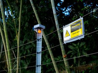 Electrical fence warning sign with electric shock icon hanging on wire and red light bulb on poles...