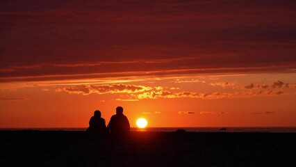 Back view of unrecognizable romantic young couple sitting on shore and enjoying stunning sunset . sea and sky, horizon. Travel, honeymoon destination