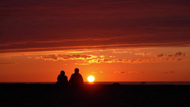 Back view of unrecognizable romantic young couple sitting on shore and enjoying stunning sunset . sea and sky, horizon. Travel, honeymoon destination