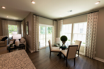 Sunny dining room with large windows, wooden upholstered chairs, floral centerpiece on a modern glass table. Real estate concept.