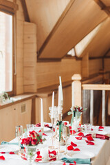 The table for a romantic dinner is decorated with red roses in vases and candles, as well as petals.