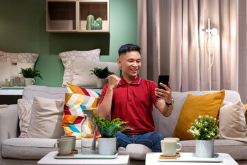 Happy Asian Man in Red Polo Shirt and Jeans Celebrating Success while looking at his Smartphone
