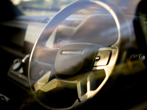 Gaydon, Warwick, UK June 2024: New Land Rover Defender Detail shot through glass of dashboard with infotainment system, steering wheel with buttons, defender logo and instrument cluster, golden hour