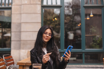 Happy young brunette woman customer shopper holding credit card using cell phone mobile app buying fashion clothes paying online make purchase in ecommerce digital store on smartphone in cafe.