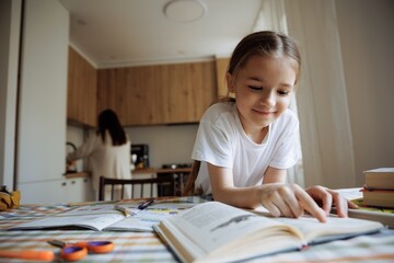 A 6-year-old daughter reads a book and does her homework against the background of her mother...