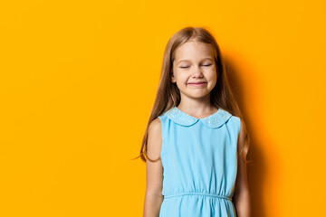 Little girl with closed eyes and a joyful smile standing against a vibrant orange wall on a sunny...