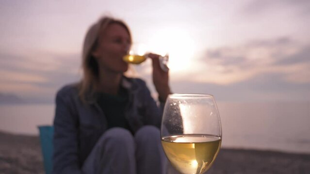 Young woman is sitting on seashore and eating delicious dough pie, holding it in her hands, and next to her are two bottles of wine. Picnic on seashore Pleasures on beach: tourist watching seascape