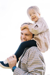 Little laughing girl hugs her smiling father head while sitting on his shoulders. Side view