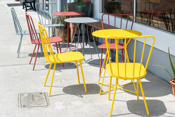 Multiple empty bright grey, yellow, and red colored metal round tables and highback chairs at a bistro cafe on a restaurant's sidewalk patio. The restaurant has large glass windows and a white wall. 