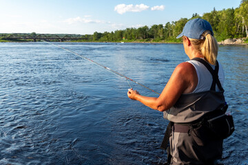 A middle age female stands in a large salmon river casting a fishing rod holding line in her left...