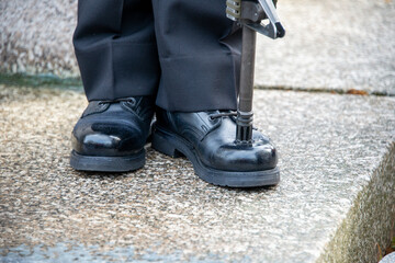 A  military person stands at attention with his rifle pitched on the toe of his shinned boot. The...