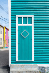 Fototapeta premium A vibrant teal blue wooden door with white trim. There's a small triangle shaped window shutter with a metal latch on the corner of the clapboard building with concrete steps to the sidewalk. 