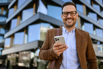 Portrait of happy male entrepreneur texting over cellphone and standing over modern office building