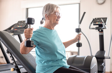 Athletic old senior woman in sportswear exercising in gym with dumbells. Elderly woman doing stretching exercise to stay fit
