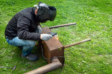 A professional welder in a protective mask welds a metal structure outdoors. Call a specialist to...