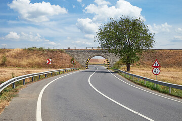 A curved rural road leading under a stone railway bridge, flanked by guardrails and road signs. A lone tree stands to the right, with a clear blue sky and scattered clouds overhead.