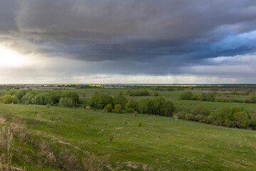 A vast, tranquil rural landscape stretches out under a dramatic sky filled with storm clouds, casting a mix of sunlight and shadows over the lush green fields and distant hills