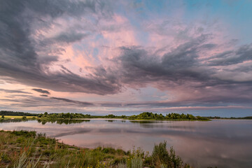 A picturesque landscape featuring a calm lake reflecting the soft hues of a spring sunset. The sky...