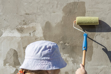 A woman in a blue vest, blue shirt and hat primes a textured concrete wall with a roller and brush....