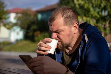 A man sits at a table outdoors, enjoying a cup of coffee while checking his phone. Hes wearing a...