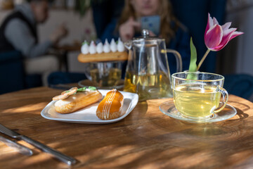 Enjoy a serene afternoon tea setting with a variety of pastries, including eclairs, served on a wooden table bathed in sunlight. The ambiance is complemented by a vibrant tulip