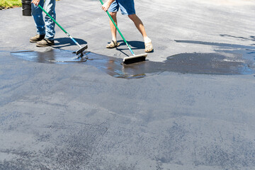 Two men applying sealer on asphalt driveway with brush and squeegee on sunny day