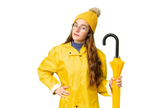 Teenager girl with rainproof coat and umbrella over isolated chroma key background posing with arms at hip and smiling
