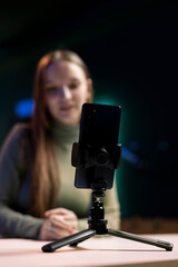 Focus on smartphone on tripod used by cheerful girl in blurry background to film in living room used as professional studio. Close up shot of phone used by teenager to record vlog
