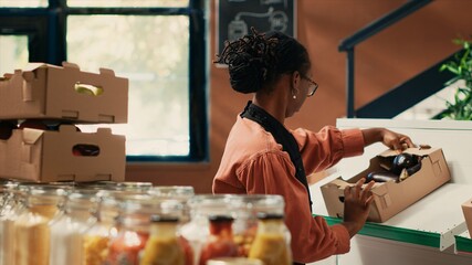 Fototapeta premium Merchant organizing fruits and veggies in crates on shelves, preparing fresh merchandise at farmers market before opening store. African american woman arranging natural produce.