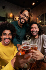Vertical portrait of multiracial group of three men friends looking at camera while being in a bar toasting with beers, celebrating.
