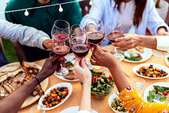 Group of young people hands toasting red wine at rooftop party. Multiracial friends celebrating at bbq dinner at home garden.