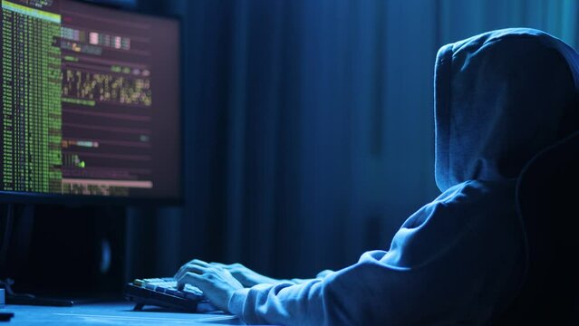 In a dimly lit room, a cloaked figure types code on a computer, hinting at a mysterious hacker